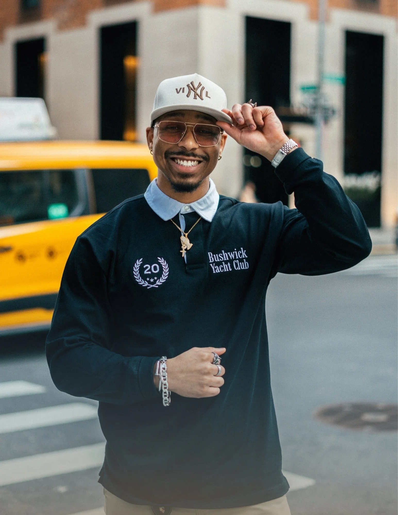 A smiling man wearing glasses, a baseball cap, a black sweatshirt with "Bushwick Yacht Club" on it, and accessories, standing on a city street with yellow taxis and buildings in the background.