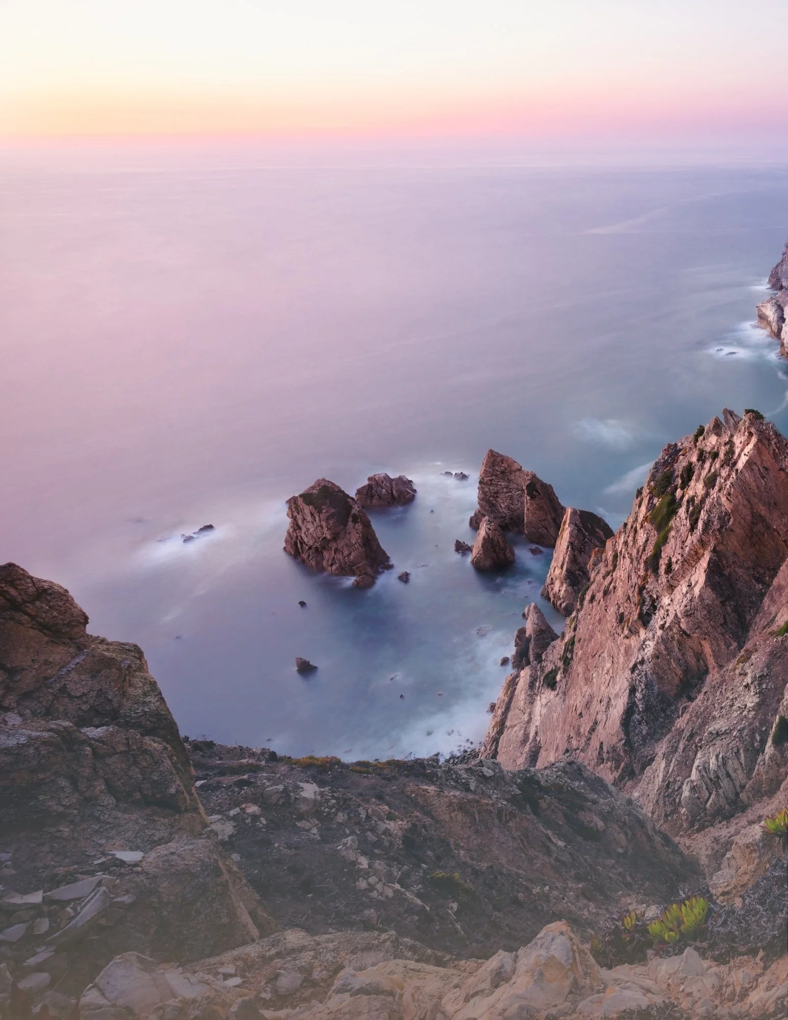 Sunset over rocky coastline with cliffs and calm ocean water in pastel pink and purple hues.
