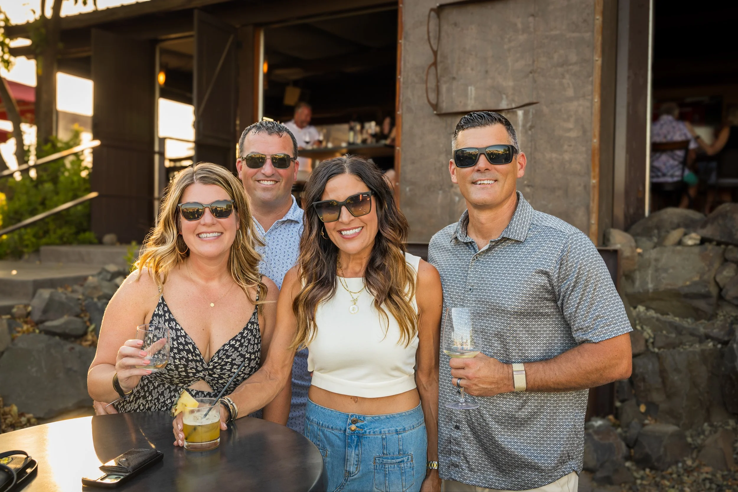 Four adults smiling, wearing sunglasses, standing outdoors at a social event, with drinks in hand, in front of a rustic wooden building or bar, during sunset.