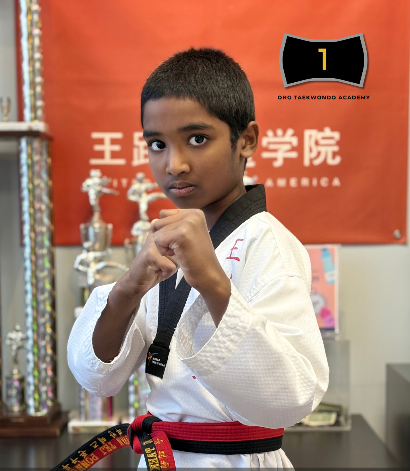 A young boy in a martial arts uniform with a black belt, standing in a martial arts studio with trophies and a red banner in the background.