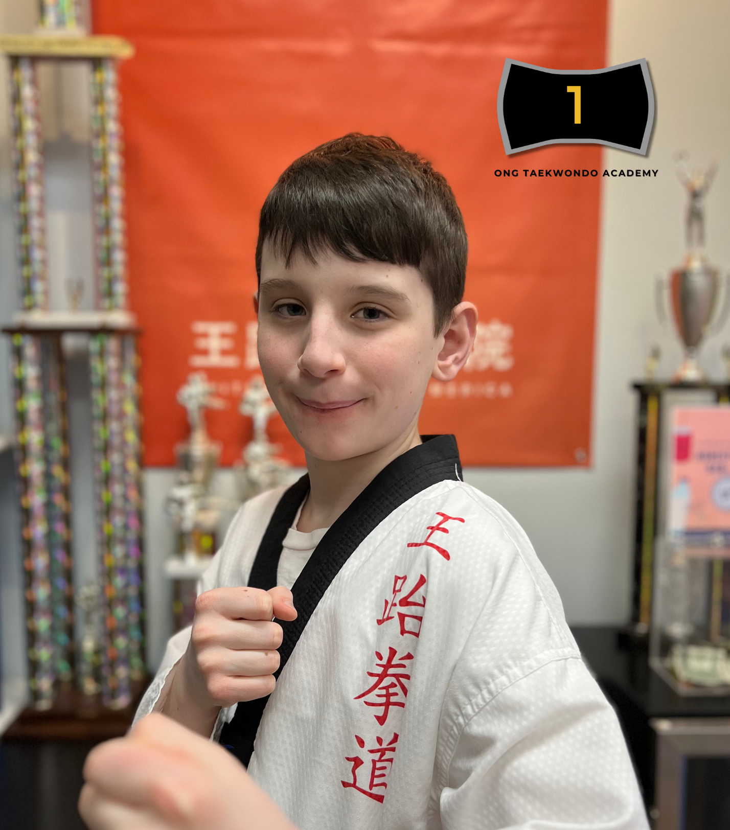 Young boy in a white martial arts gi with red character embroidery, posing with a fist at a taekwondo academy with trophies in the background.