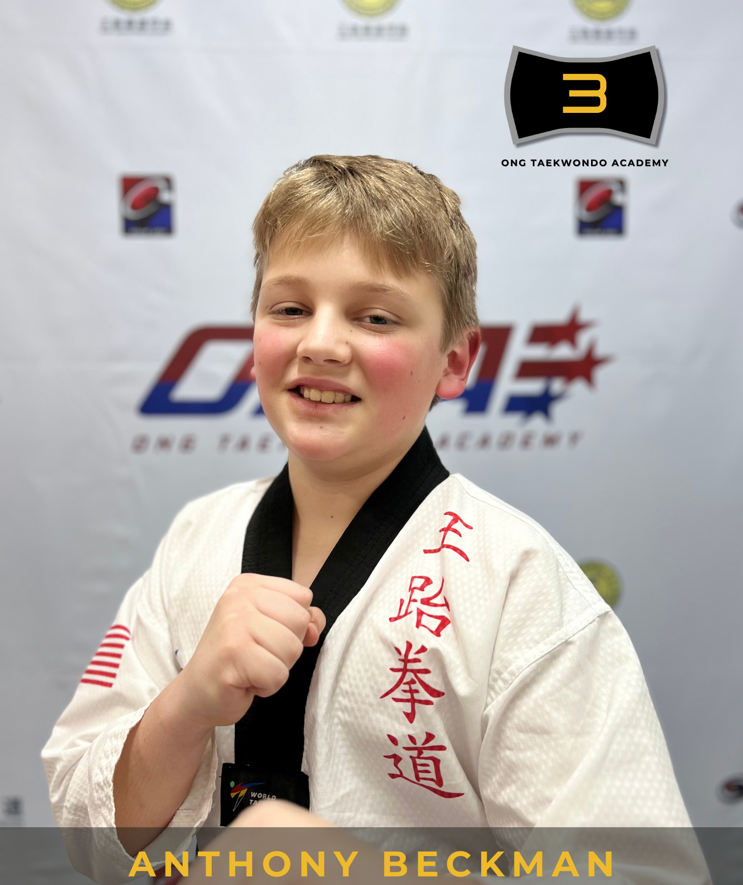 A young boy in a white Taekwondo uniform with red embroidery on the right side, standing in front of a backdrop with various logos, showing a black belt and smiling while holding his fist in a martial arts pose.