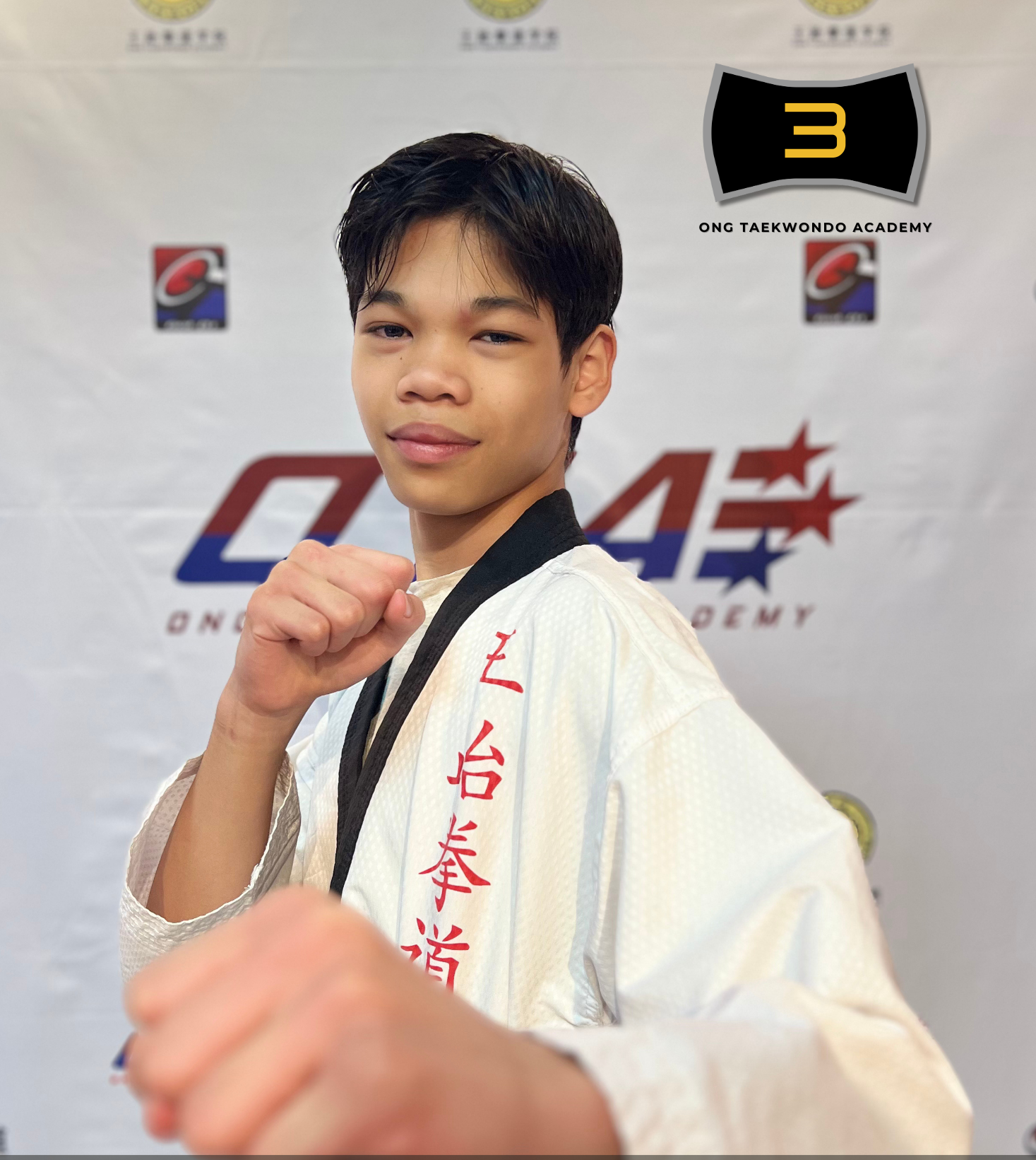 Young martial artist in a white gi with red Asian characters, holding a fist forward, standing in front of a backdrop with logos, celebrating earning a black belt at a martial arts academy in 2024.