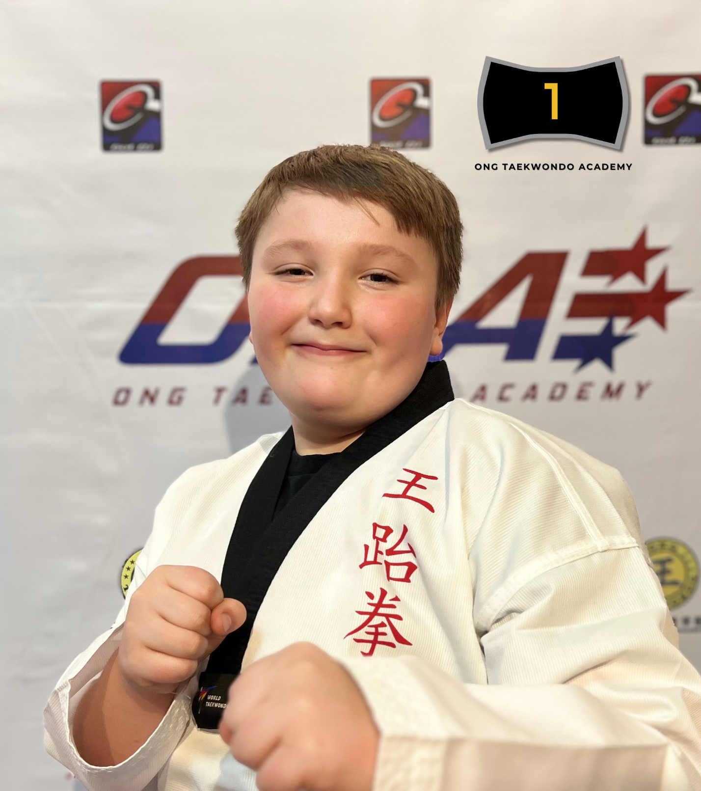 A young boy in a martial arts uniform posing with a smile and a clenched fist, standing in front of a backdrop with logos of the ONG Taekwondo Academy, indicating he has received a black belt in 2024.