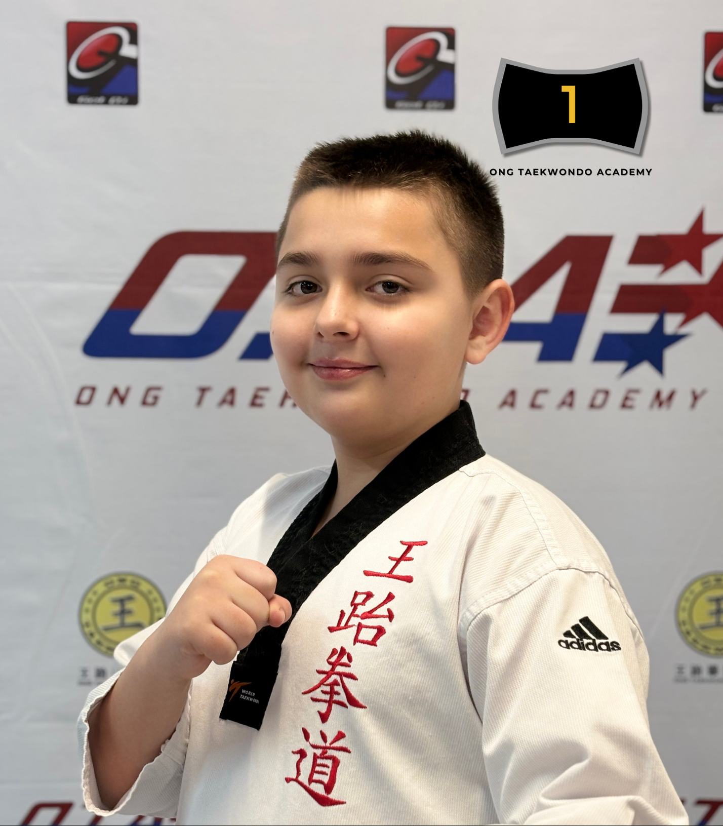 A young boy in a taekwondo uniform with red Japanese kanji on the chest, standing in front of a backdrop with logos for O.G. Taekwondo Academy, holding a black belt with his right hand in a fist, celebrating his achievement for the year 2022.