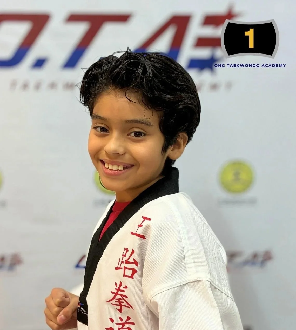 Young boy smiling, wearing a taekwondo uniform and black belt, standing in front of a taekwondo academy banner.