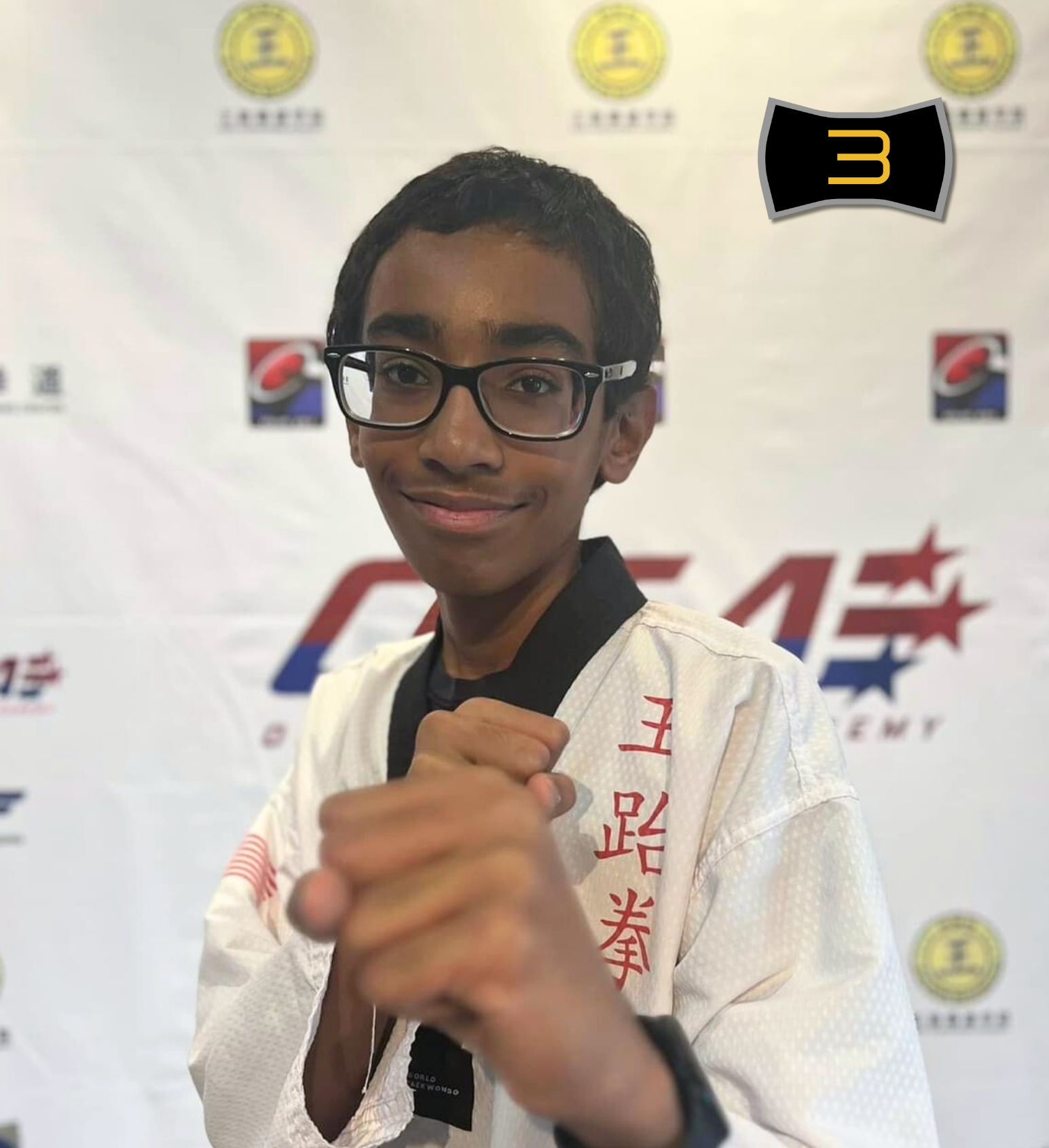 Young male taekwondo student wearing a white gi, black belt, and glasses, making a fist in front of a Taekwondo academy backdrop with logos and text that says 'Ong Taekwondo Academy Instructor Batch 5'.