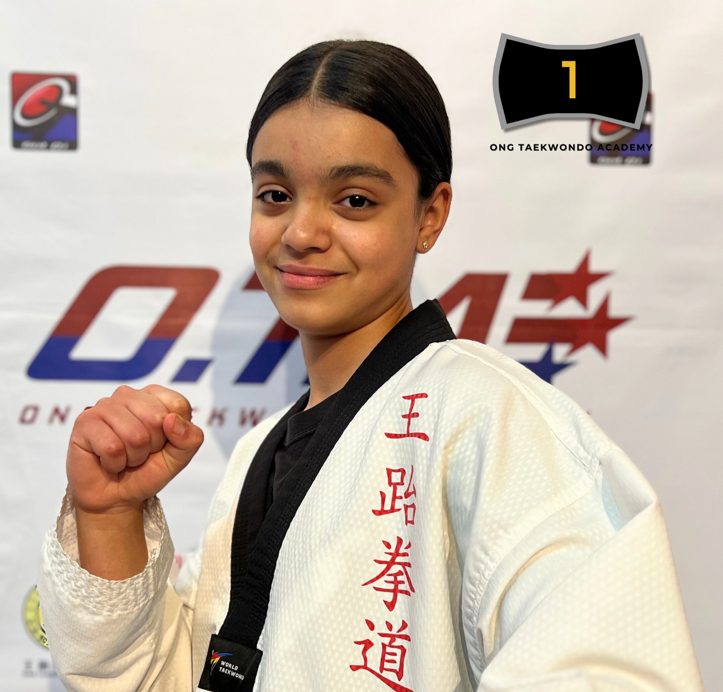 Young female martial artist wearing a white gi with red Korean characters, black belt, and fist raised in a fighting stance, standing in front of a backdrop with logos and text related to taekwondo.