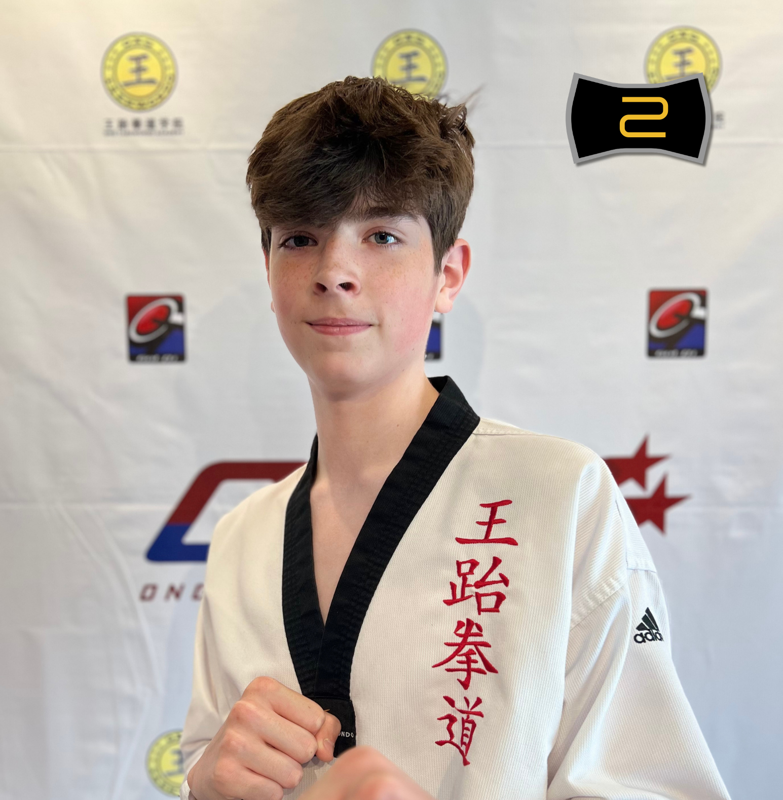 Young male martial artist in a white taekwondo uniform with black collar, holding his fist up, standing in front of a banner with logos and medals, indicating he is an instructor at ONG Taekwondo Academy, Batch 5.