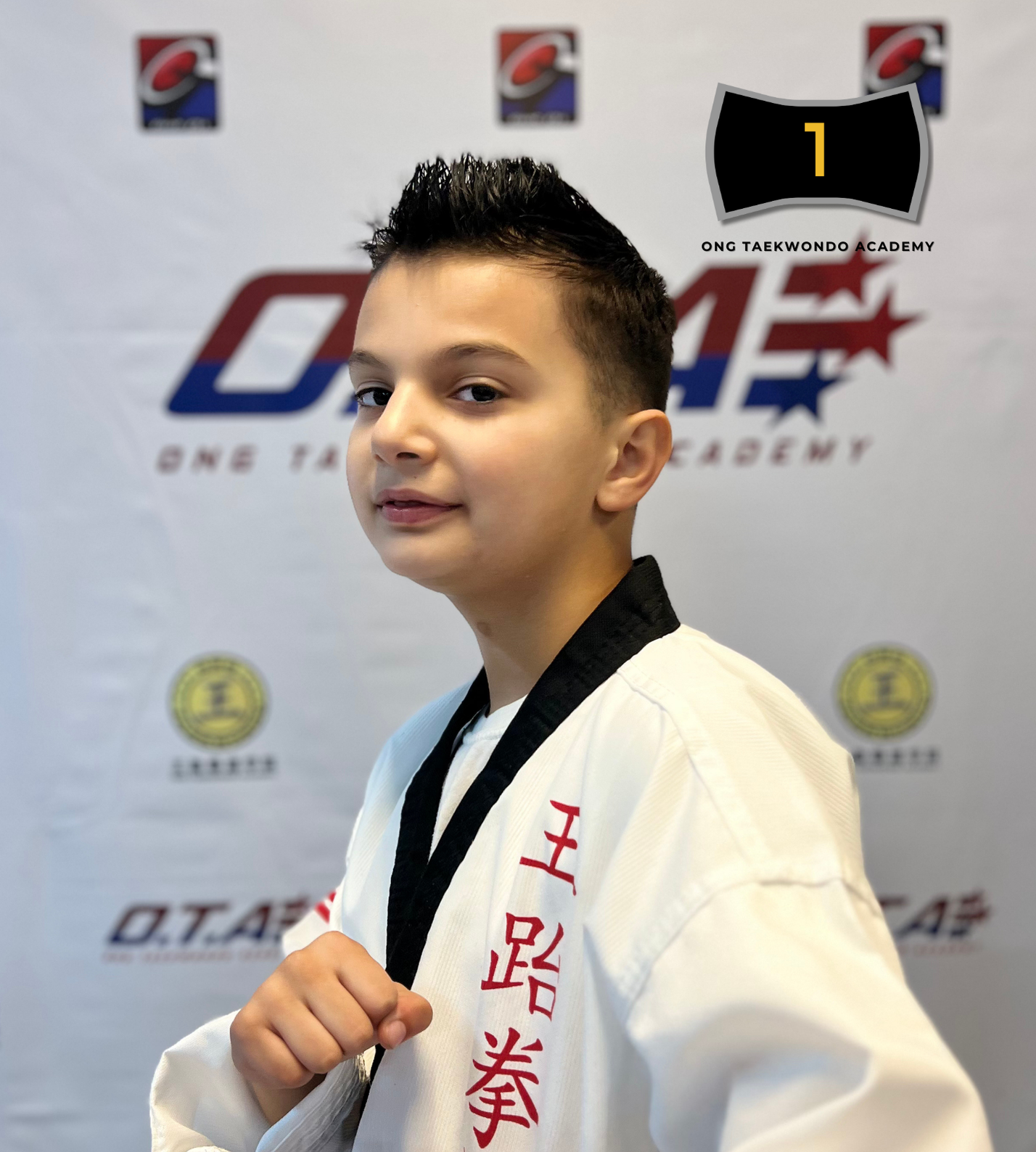 Young boy wearing a white taekwondo uniform with red Chinese characters, posing in front of a taekwondo academy banner, holding his fist up in a martial arts stance.