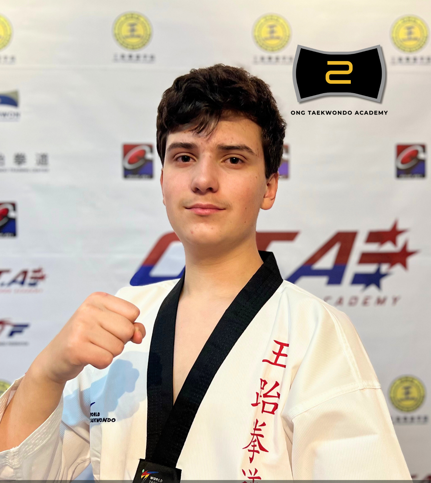 Young male martial artist wearing a white taekwondo uniform with red writing, holding a black belt and making a fist, standing in front of a backdrop with logos and the words 'Ong Taekwondo Academy.'