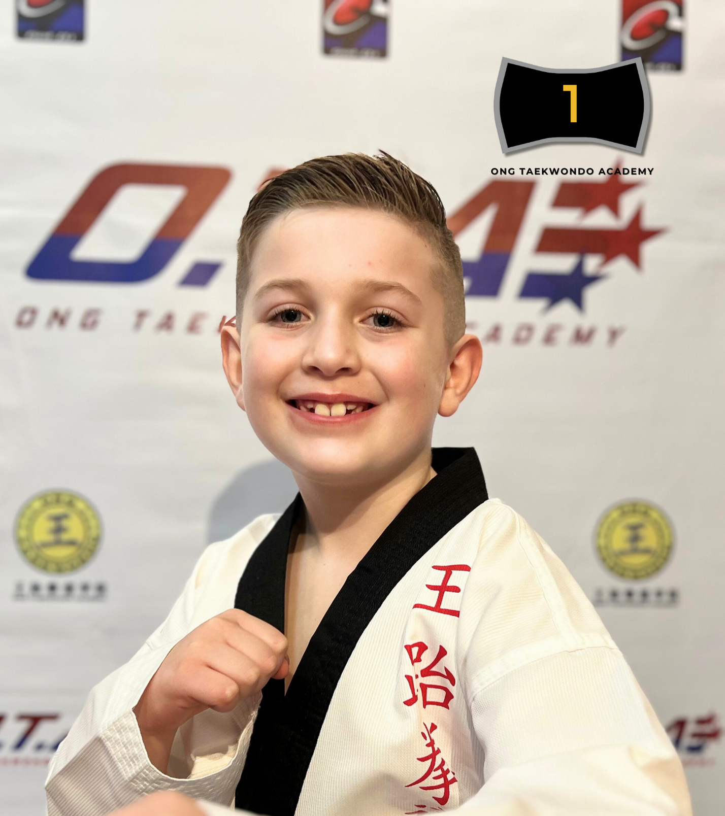 Young boy with short, slicked-back hair and a wide smile, wearing a white taekwondo uniform with black collar and red characters on the left side, posing in front of a backdrop with taekwondo logos and banners, holding a fist up. Text overlay reads: "Greyson Rettele Black Belt Year 2024."