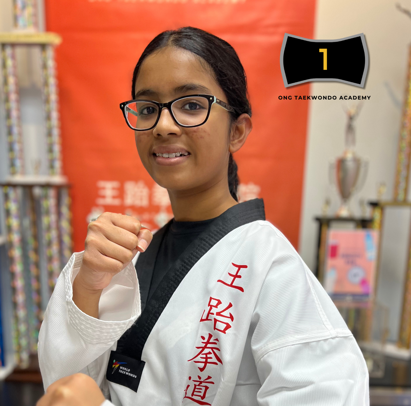 A young woman wearing glasses in a martial arts uniform with red Chinese characters on it, standing in a martial arts academy with trophies and trophies in the background, making a fighting stance.