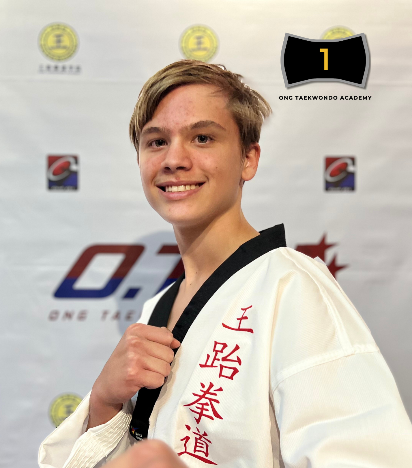 Young man with a black belt in martial arts, wearing a white gi with red Chinese characters, smiling and holding his fist up, standing in front of a backdrop with various logos and text.