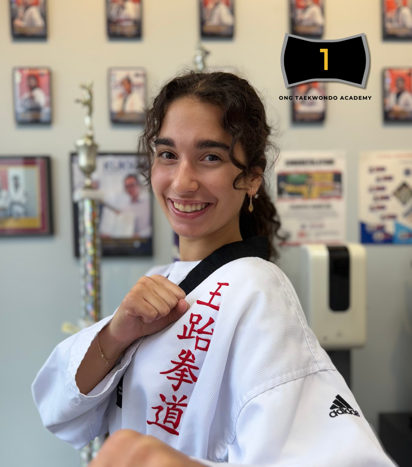 Smiling young woman in a white taekwondo uniform with red Japanese characters, holding a black belt over her shoulder, in a martial arts studio with trophies and posters on the wall.