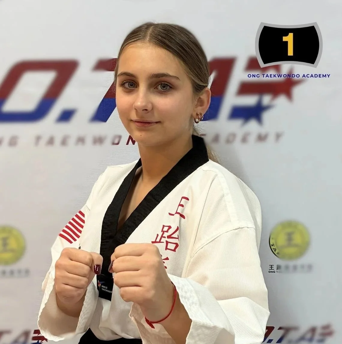 Young woman in a white martial arts uniform with black collar, standing in front of a background with logos. She is holding her fists up in a fighting stance.