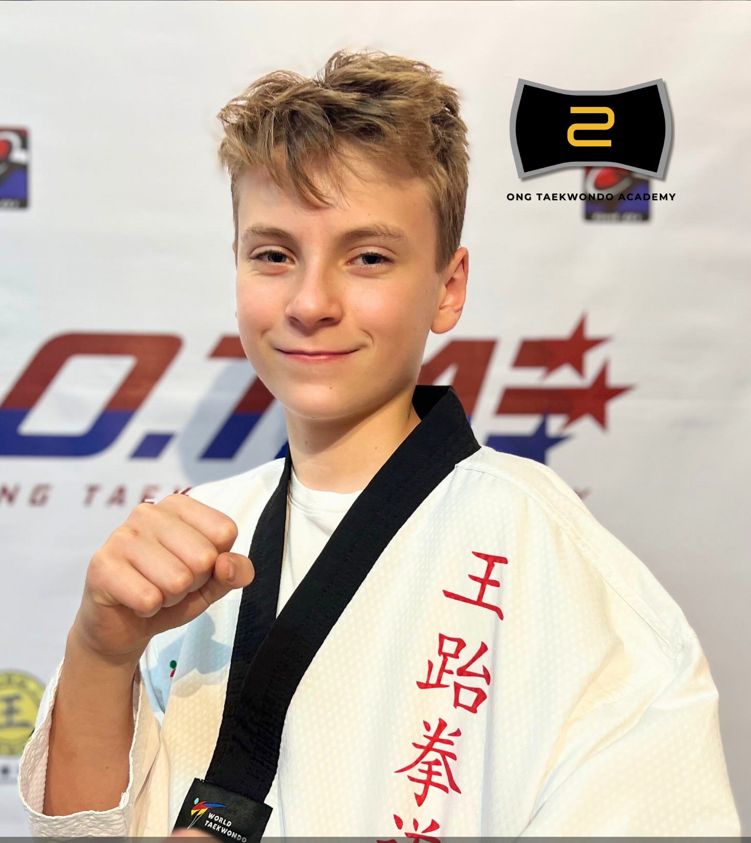Young male martial artist wearing a white judo gi with red Japanese characters on the sleeve, holding a black belt, smiling at the camera, with a backdrop featuring logos related to taekwondo.