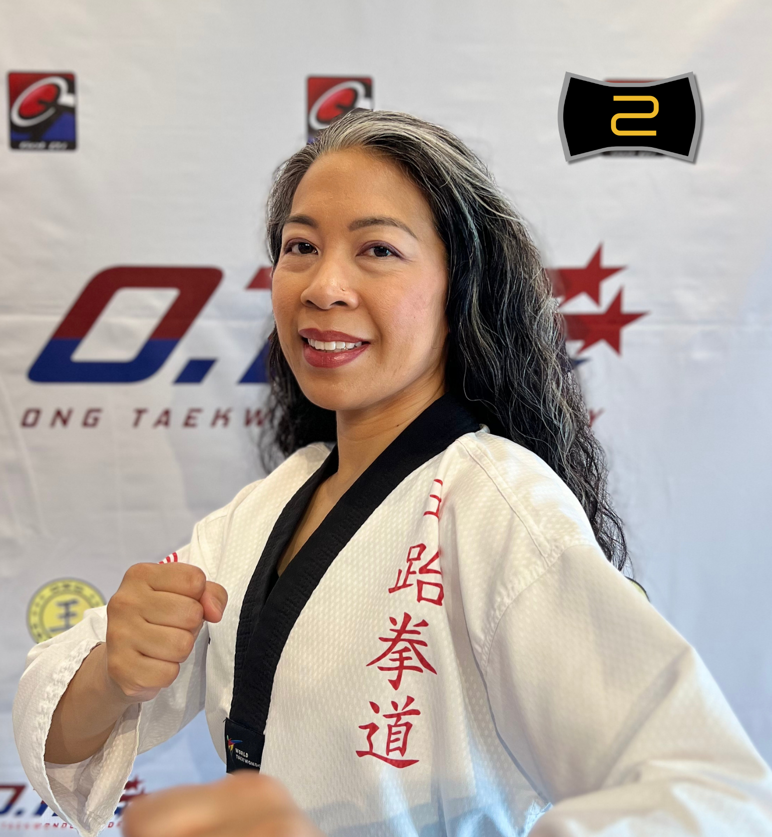 Portrait of a woman dressed in a white karate uniform with red Japanese characters, smiling and posing with a fist in front of a banner with logo and text for ONG Taekwondo Academy, batch 5 instructor.