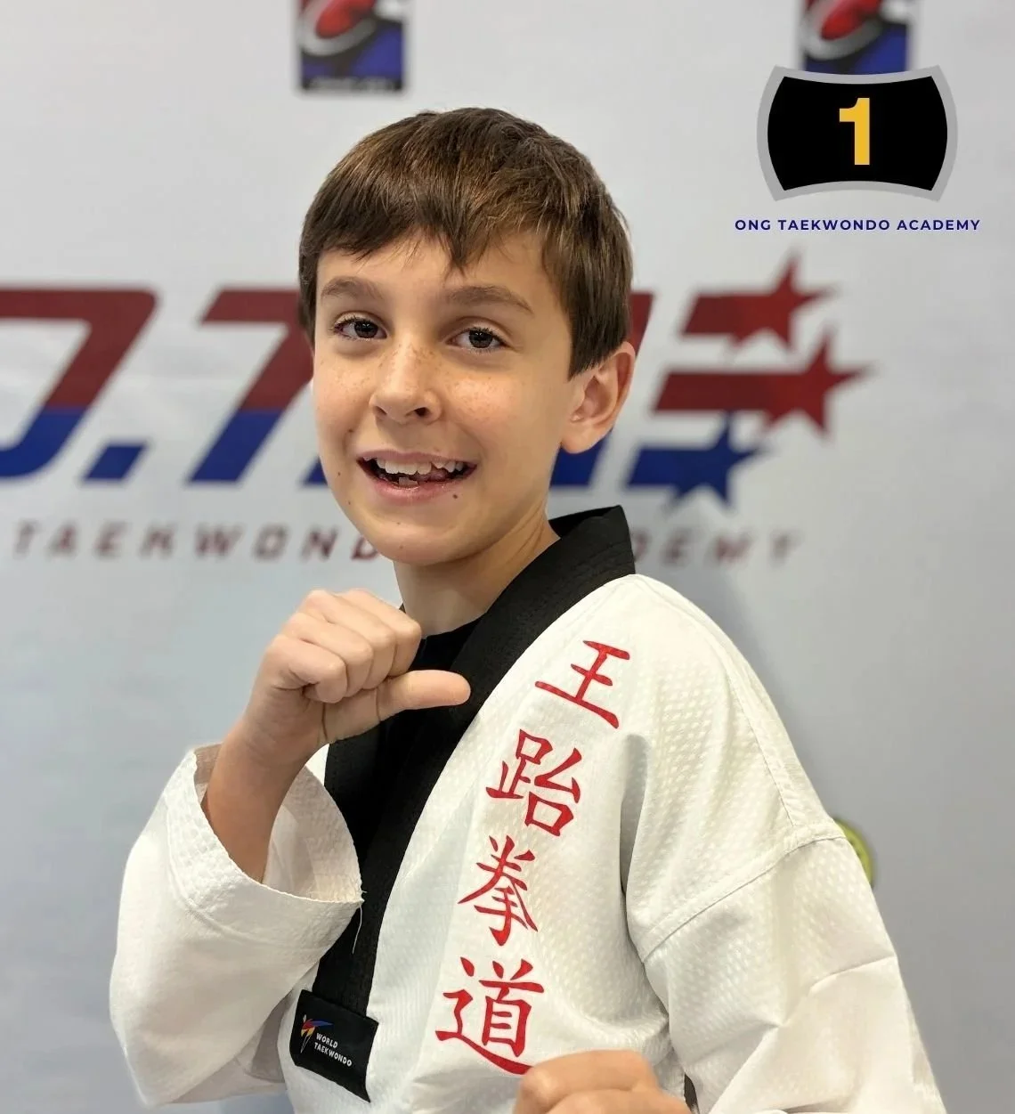 Young boy in a taekwondo uniform, smiling and holding his fist up in a martial arts pose, in front of a taekwondo academy logo.