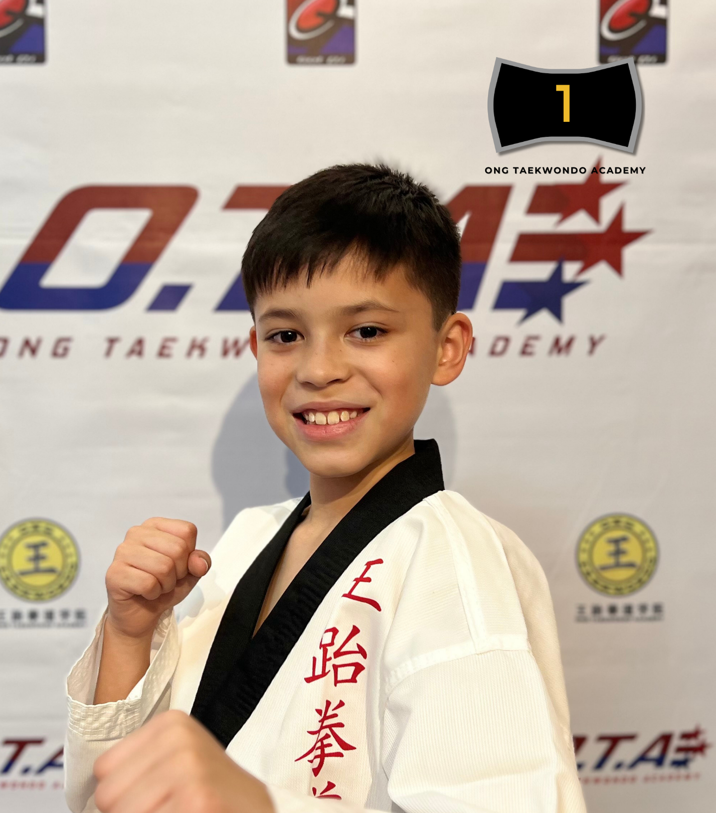 A young boy in a taekwondo uniform with a black belt, smiling and posing with a fist in front of a backdrop with taekwondo logos and text.