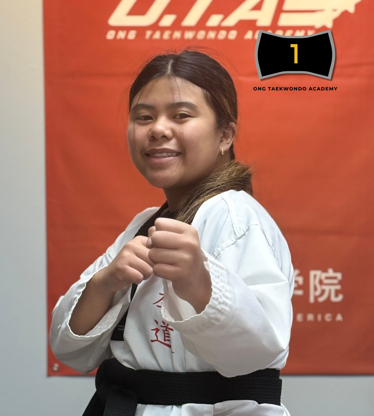 A young girl with long hair in a ponytail, wearing a white martial arts uniform with a black belt, standing in a martial arts studio. She is holding her fists up in a fighting stance, smiling confidently. Behind her is an orange banner with white and black text related to a taekwondo academy.