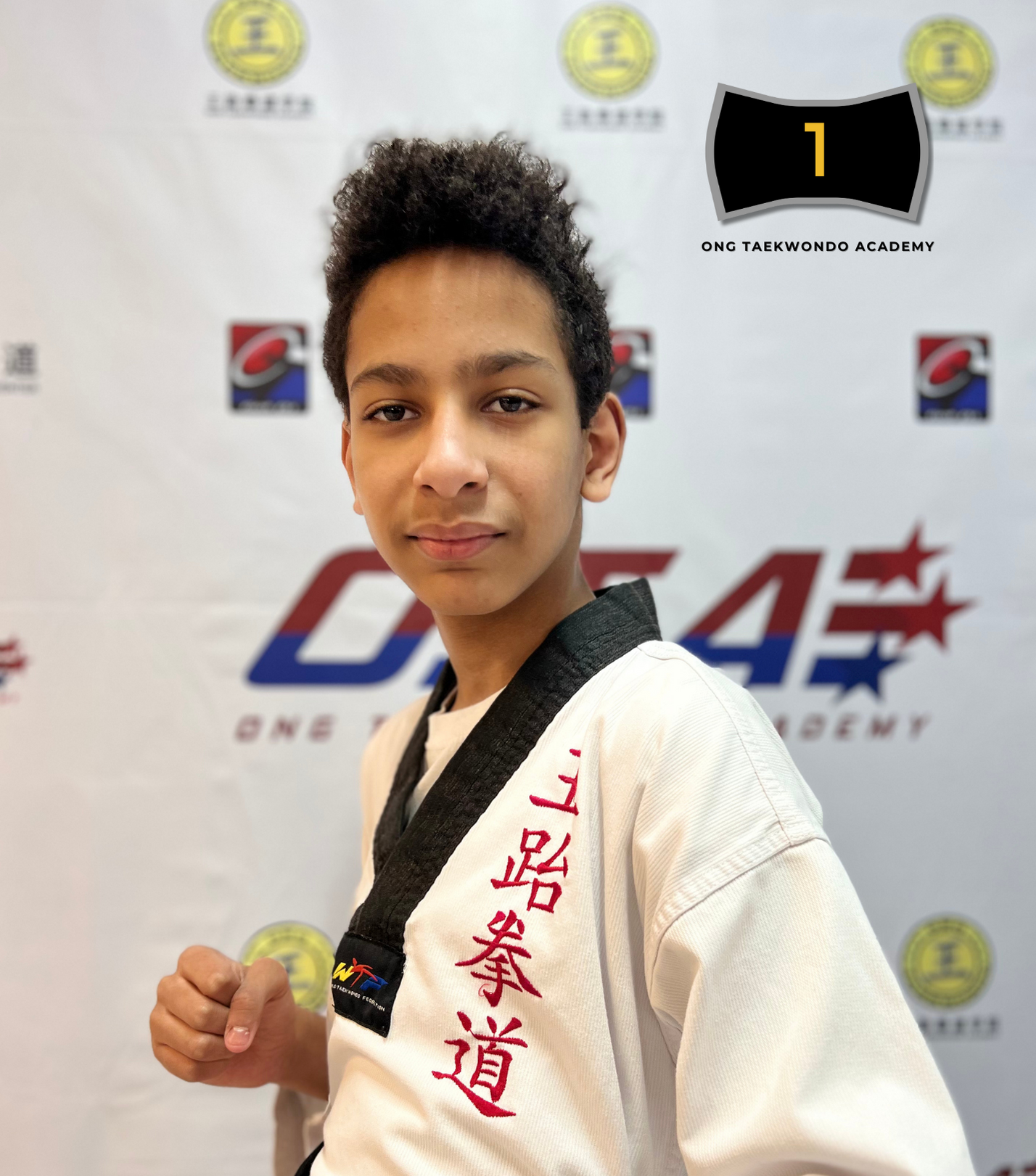 Young male martial artist wearing a black belt and white uniform with red embroidery, standing in front of a backdrop with martial arts logos and text, posing confidently with a fist.