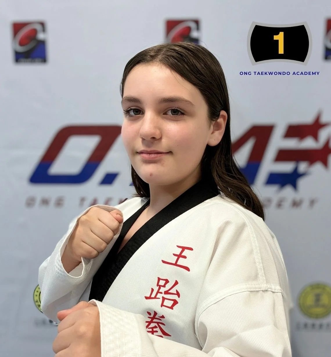 Young girl in a white taekwondo uniform with red Chinese characters, standing in front of a taekwondo logo and emblem, showing a confident stance with her fists closed.