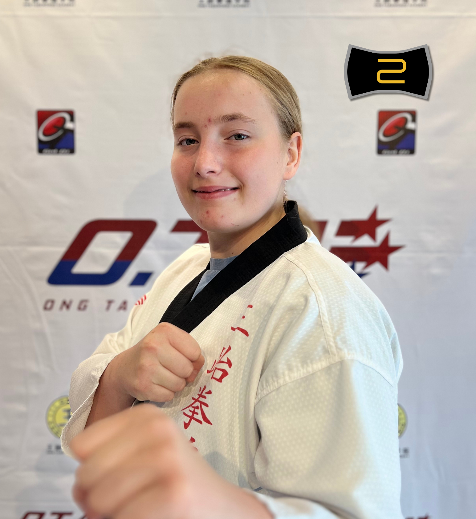 A young female taekwondo instructor in a white taekwondo uniform with black collar, posing with a martial arts stance and a confident expression, standing in front of a backdrop with logos and text related to the academy and batch 5.