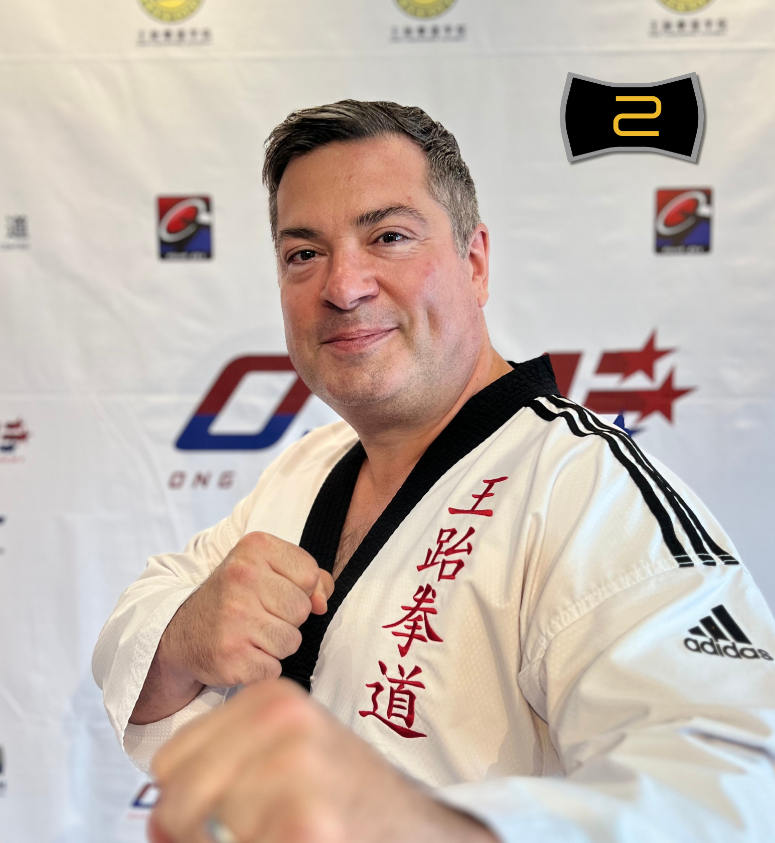 A man in a white Taekwondo uniform with black trim and red Japanese characters, posing in front of a step and repeat banner with logos.