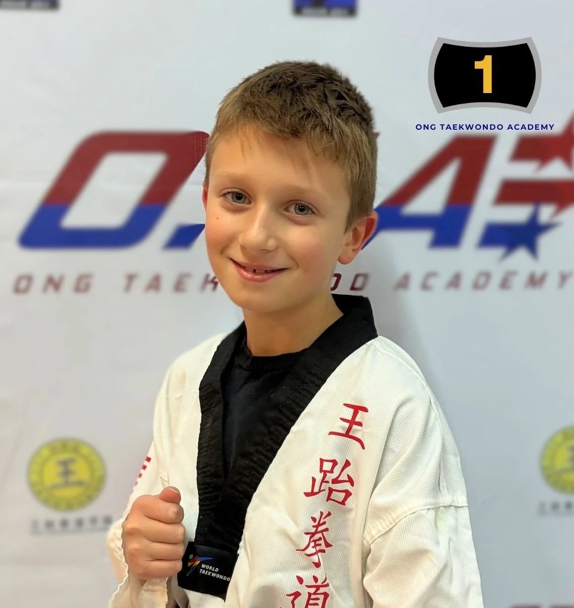 A young boy in a white martial arts uniform with red Chinese characters, smiling and holding a black belt in front of a banner that says 'Ong Taekwondo Academy' with a logo featuring a black shield and the number 1.