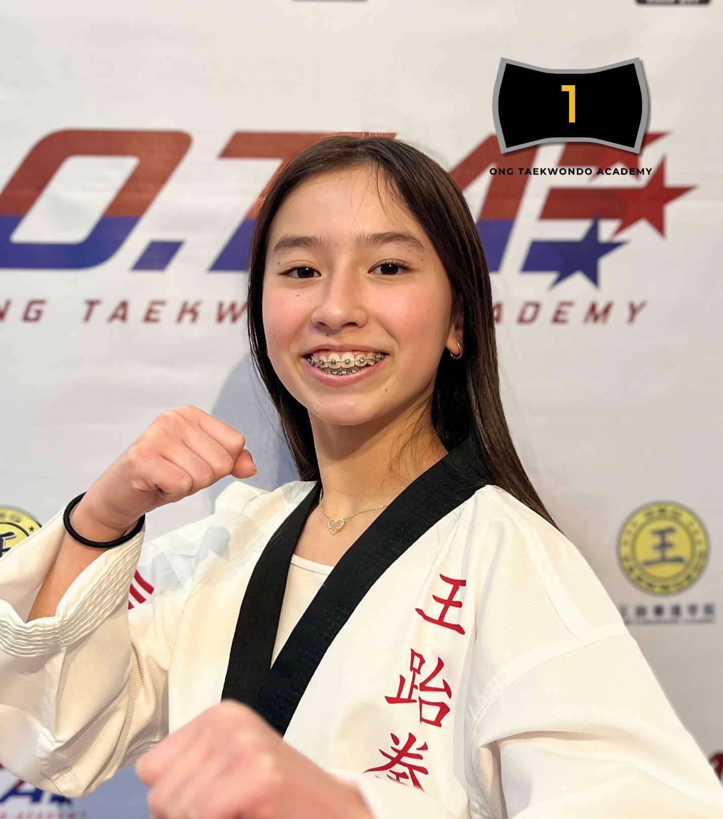 Young girl in a white taekwondo uniform with a black belt, smiling and showing a fist gesture with braces on her teeth in front of a taekwondo academy backdrop.