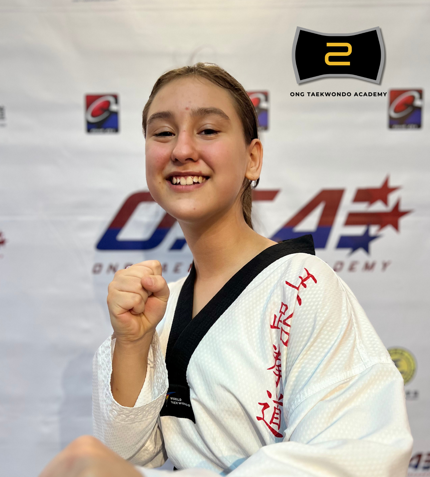 Sara Lozanski in a white taekwondo uniform with a black belt, smiling and showing her fist in front of a taekwondo academy backdrop.