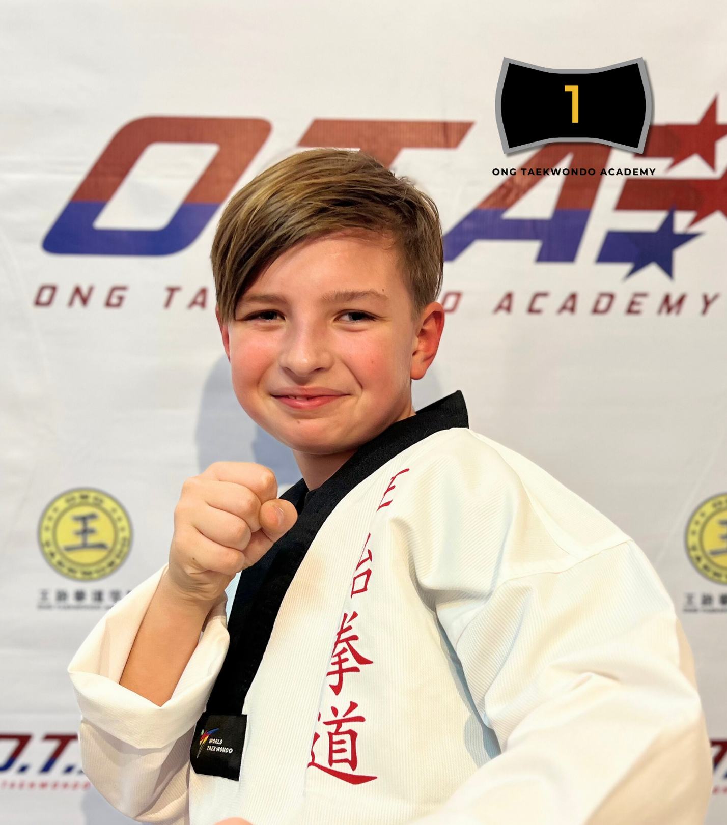 Young boy in a martial arts gi holding a fist, smiling, with a backdrop displaying 'Ong Taekwondo Academy' and a black belt label, celebrating his black belt achievement in 2024.