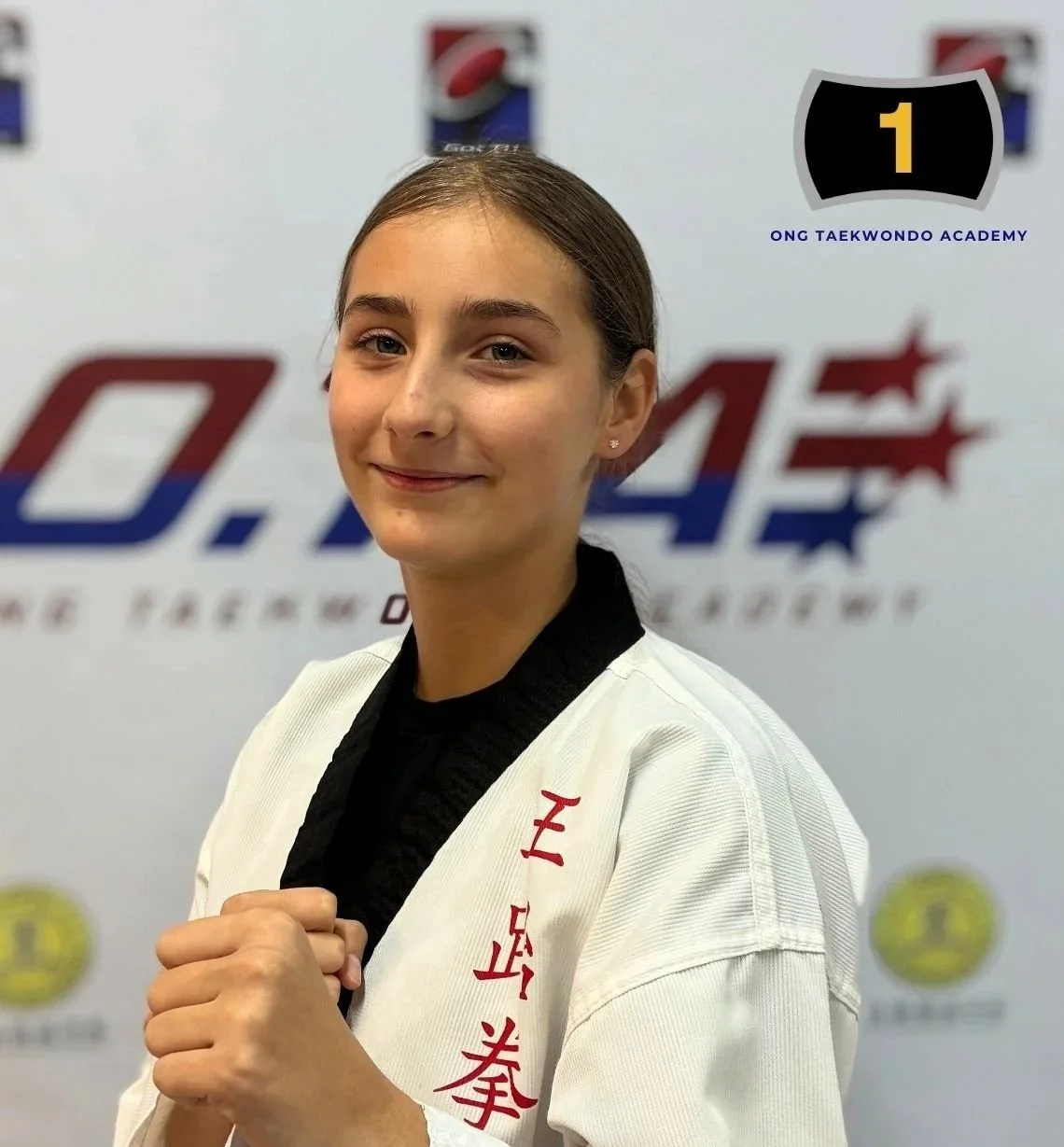 Young girl in a white taekwondo uniform smiling and posing in front of a sports banner that reads 'Q.44' and 'Ong Taekwondo Academy'.