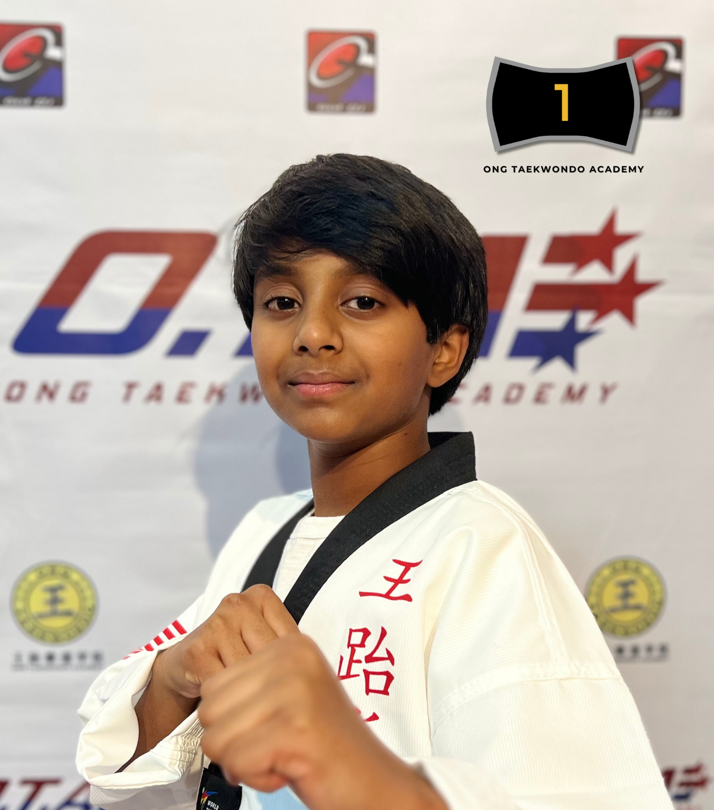 Young boy in a white martial arts gi with black belt, posing with a fist in front of a backdrop featuring the Ong Taekwondo Academy logo.