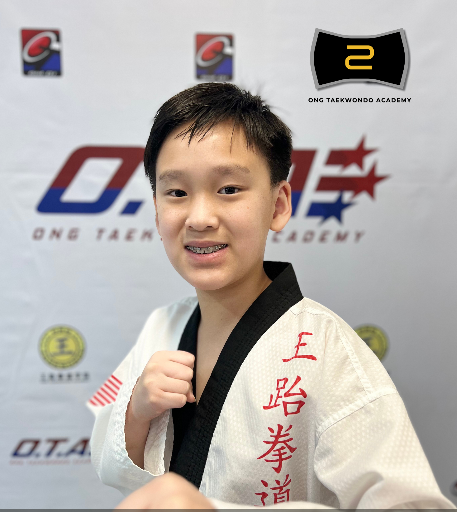Young boy wearing a white Black Belt taekwondo uniform with red Chinese characters, smiling and preparing for a taekwondo pose at a martial arts event.