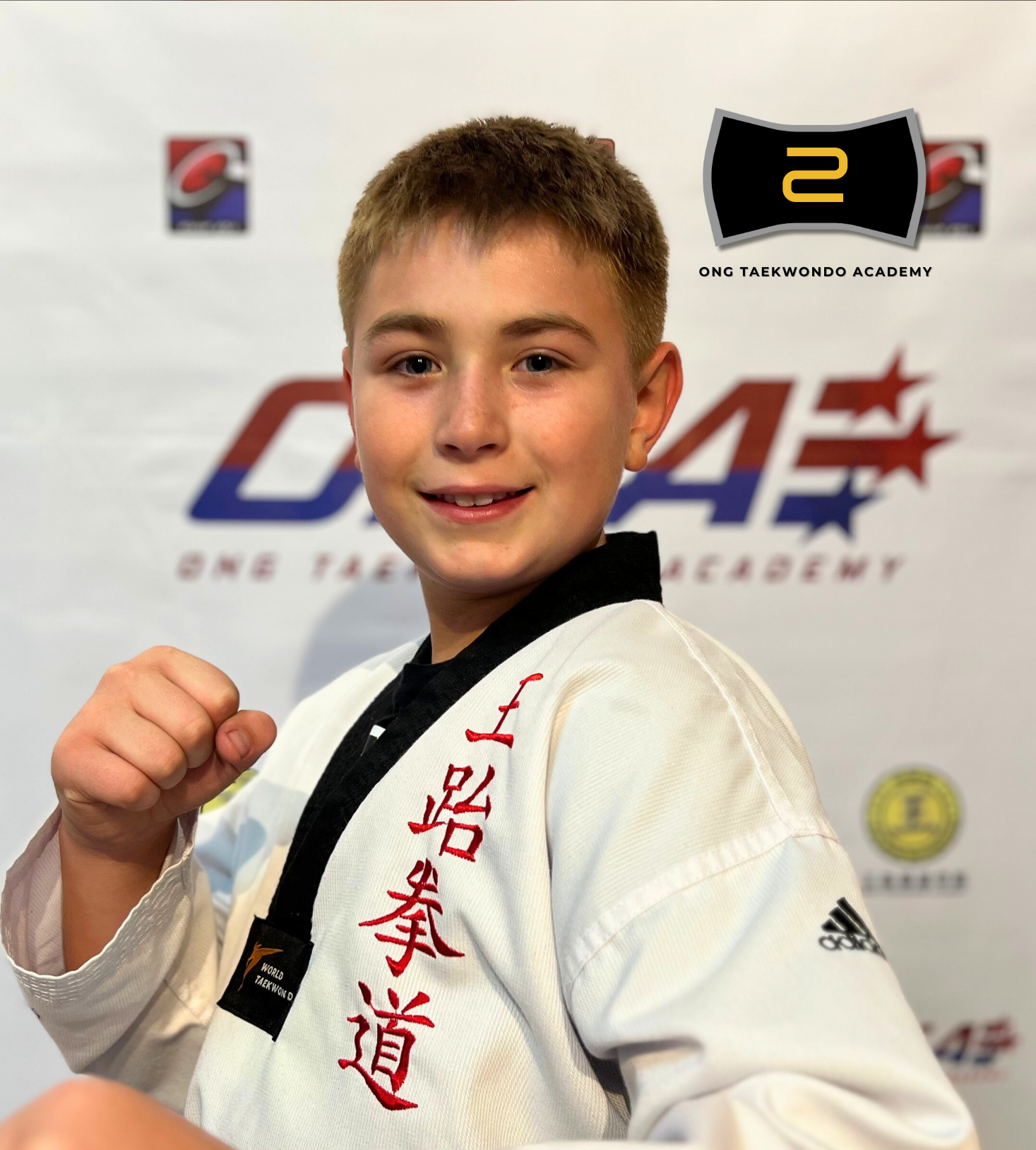A young boy in a white taekwondo uniform with red and black embroidery, posing with a fist in front of a backdrop with the logo of the Ong Taekwondo Academy and other logos, celebrating earning a black belt in 2024.