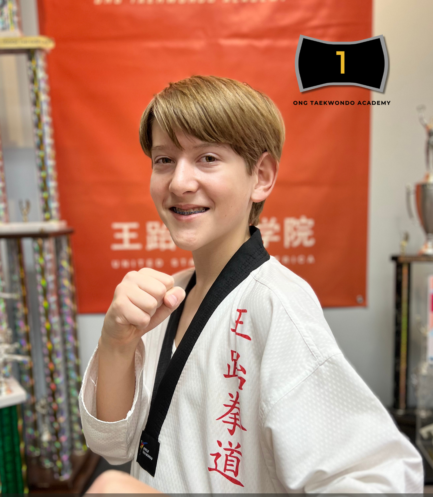 Ilia Nikonchuk in a white martial arts uniform with red Chinese characters, smiling with a fist raised, standing in a martial arts studio with trophies and a red banner in the background.