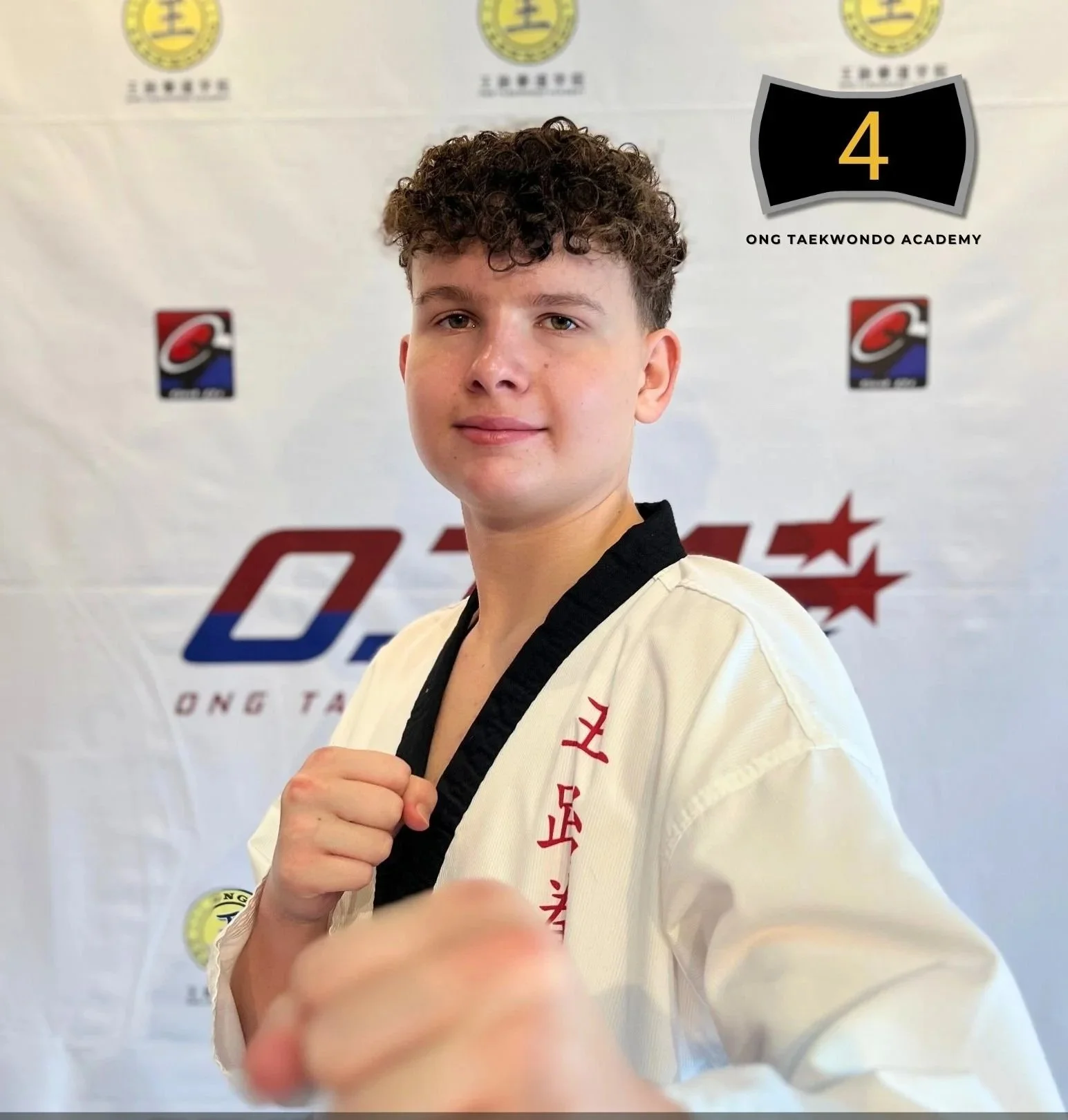 Young male martial artist in a white taekwondo uniform with black trim, standing confidently with fists raised in a fighting stance, in front of a school banner showing the number 4 and various logos.