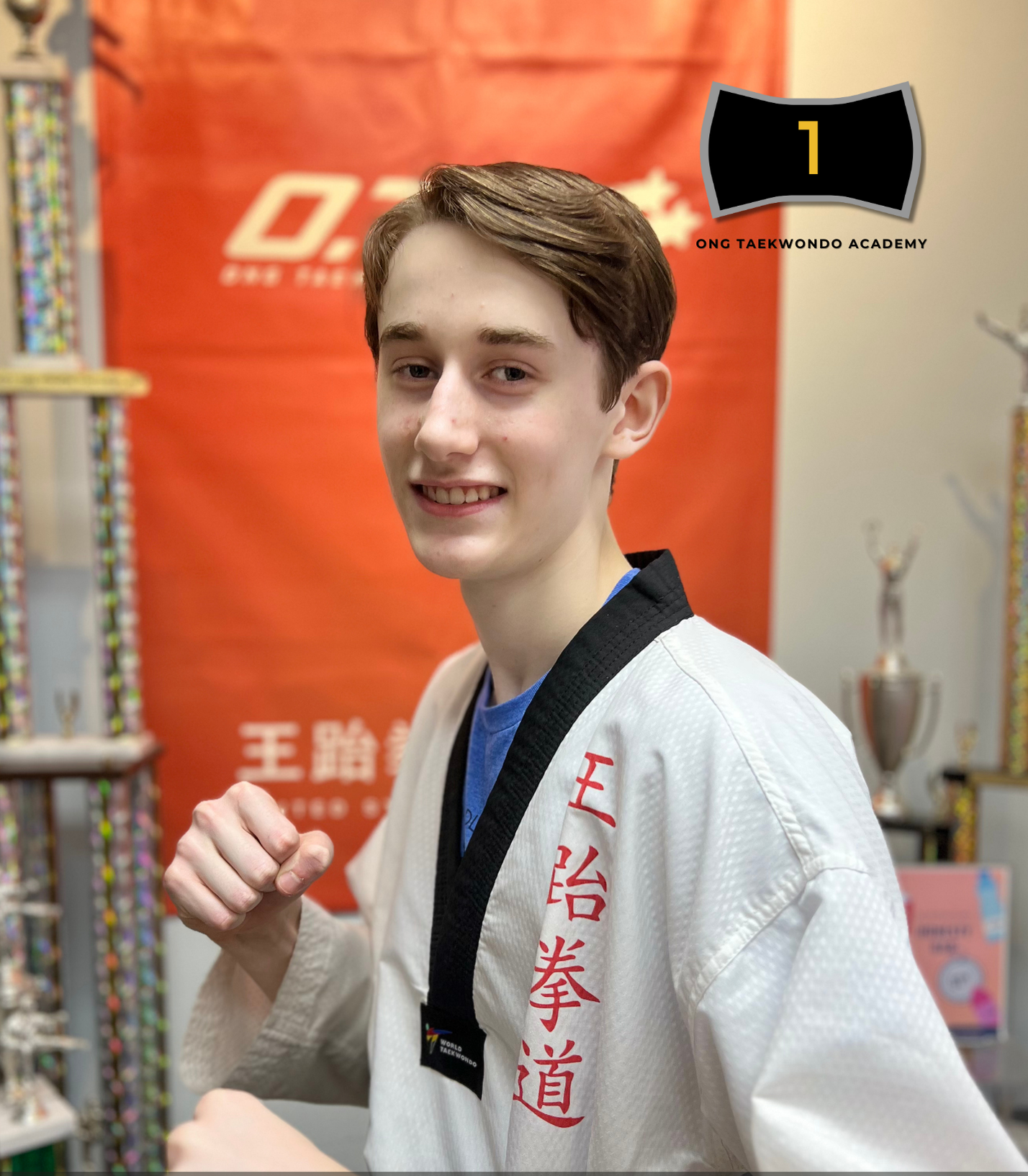 Young man wearing a white taekwondo uniform and black belt posing with a fist in front of a red backdrop, with trophies and medals in the background.