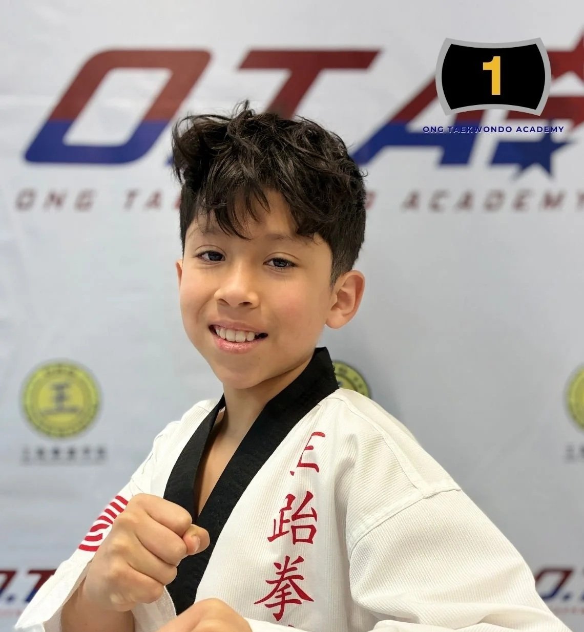 Young boy with short, dark, curly hair, smiling and holding a martial arts pose, wearing a white martial arts uniform with red and black writing, standing in front of a backdrop with logos related to Tae Kwon Do.
