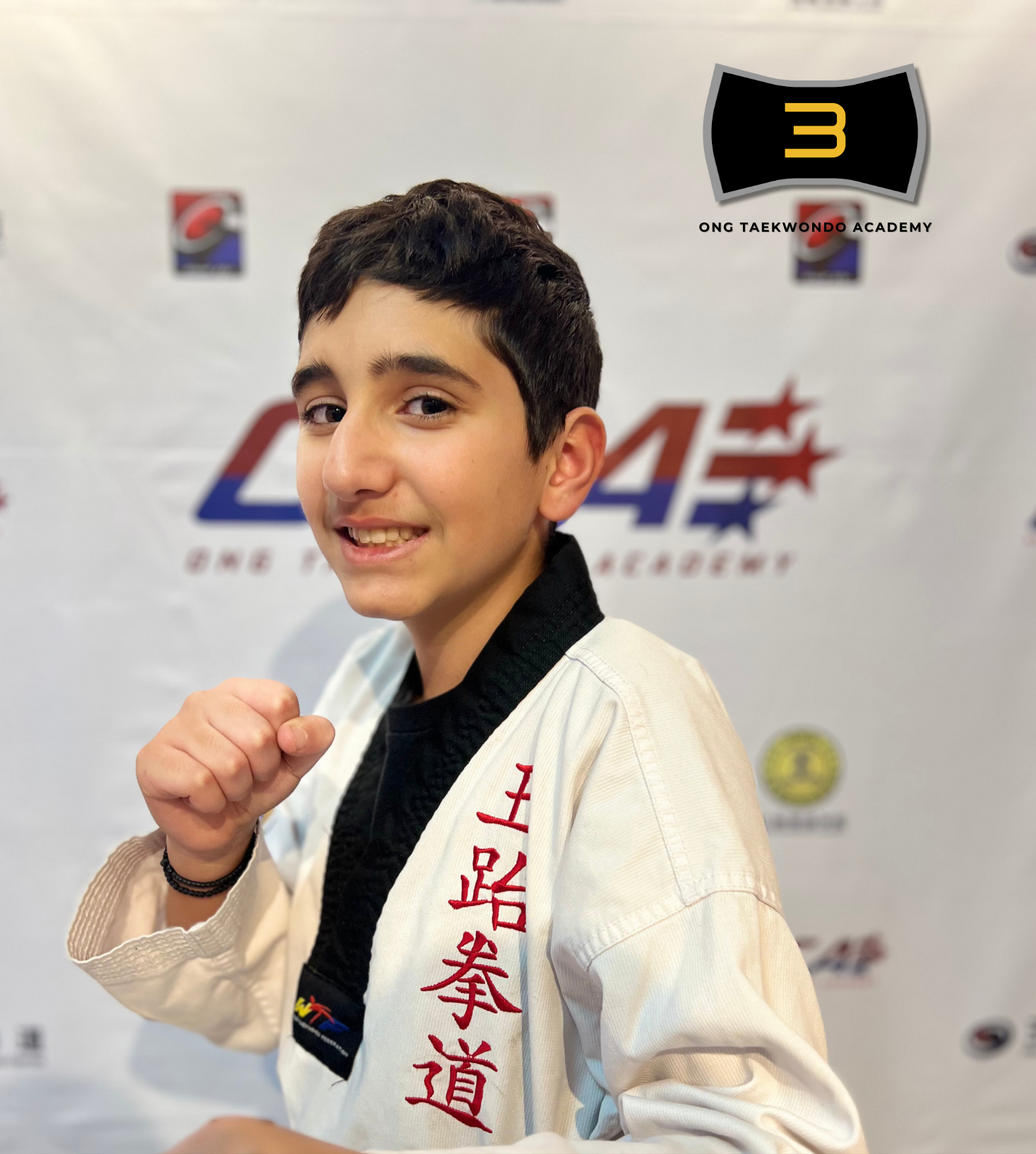 Young boy in a martial arts uniform posing with a fist raised, standing in front of a backdrop with various logos, including Ong Taekwondo Academy. Text overlay reading 'Christos Styliaras Black Belt Year 2023'.