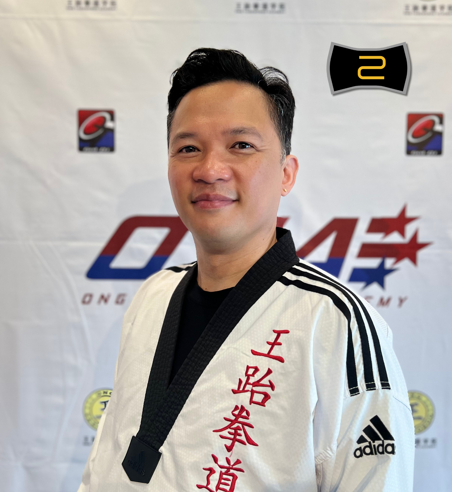 A smiling man in a white taekwondo uniform with red Chinese characters, an Adidas logo, and black trim, standing in front of a backdrop with various logos including ONG Taekwondo Academy.