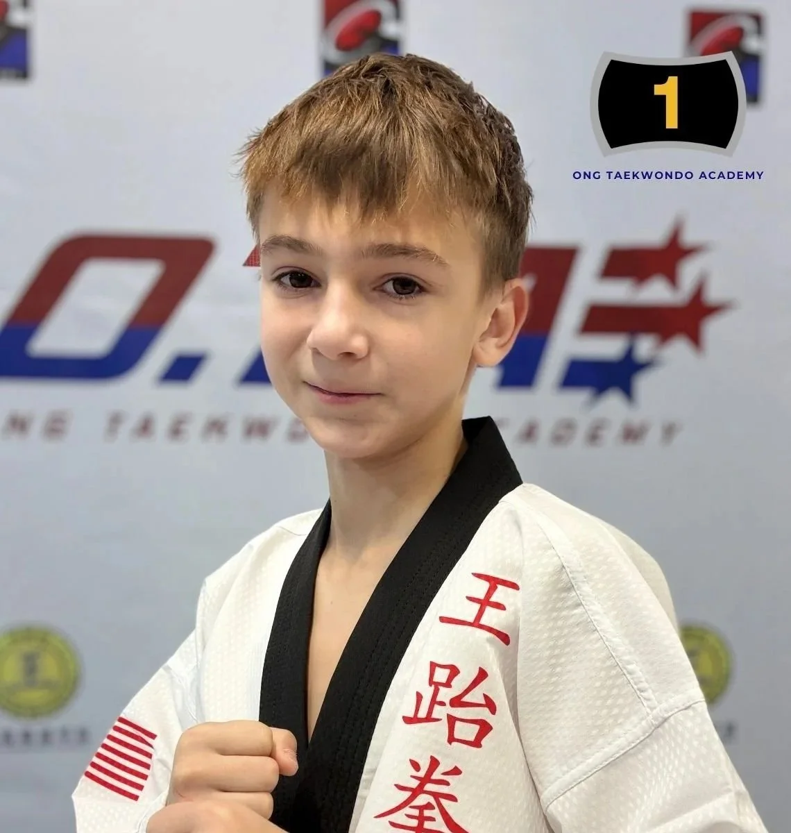 Young boy in a white taekwondo uniform with black collar, standing in front of a martial arts academy logo, holding his hand in a fist in a martial arts stance.