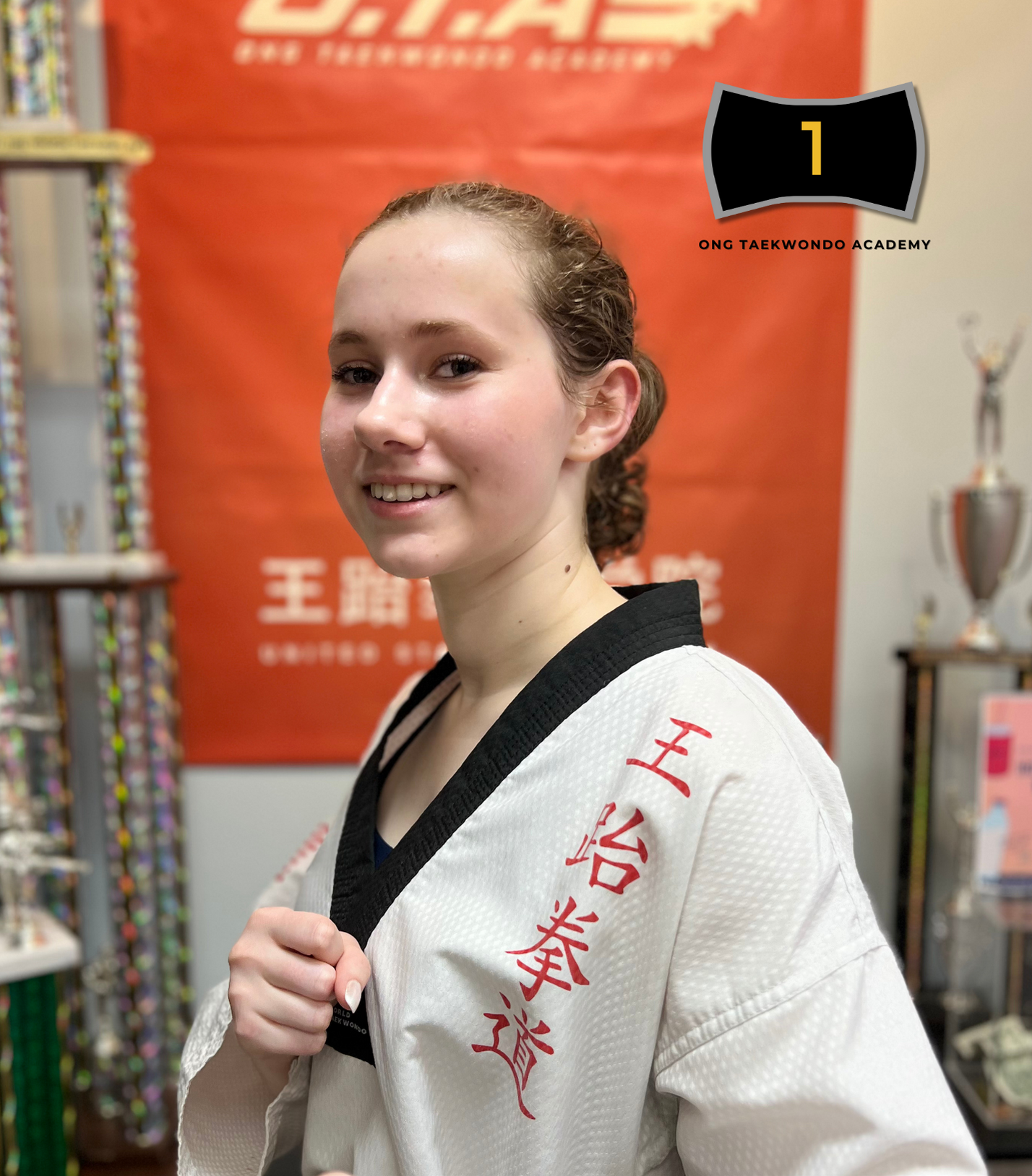 Young woman in a white taekwondo uniform with black trim, standing in a martial arts school, smiling and holding her uniform lapel. Background includes trophies, equipment, and a red banner with white text.