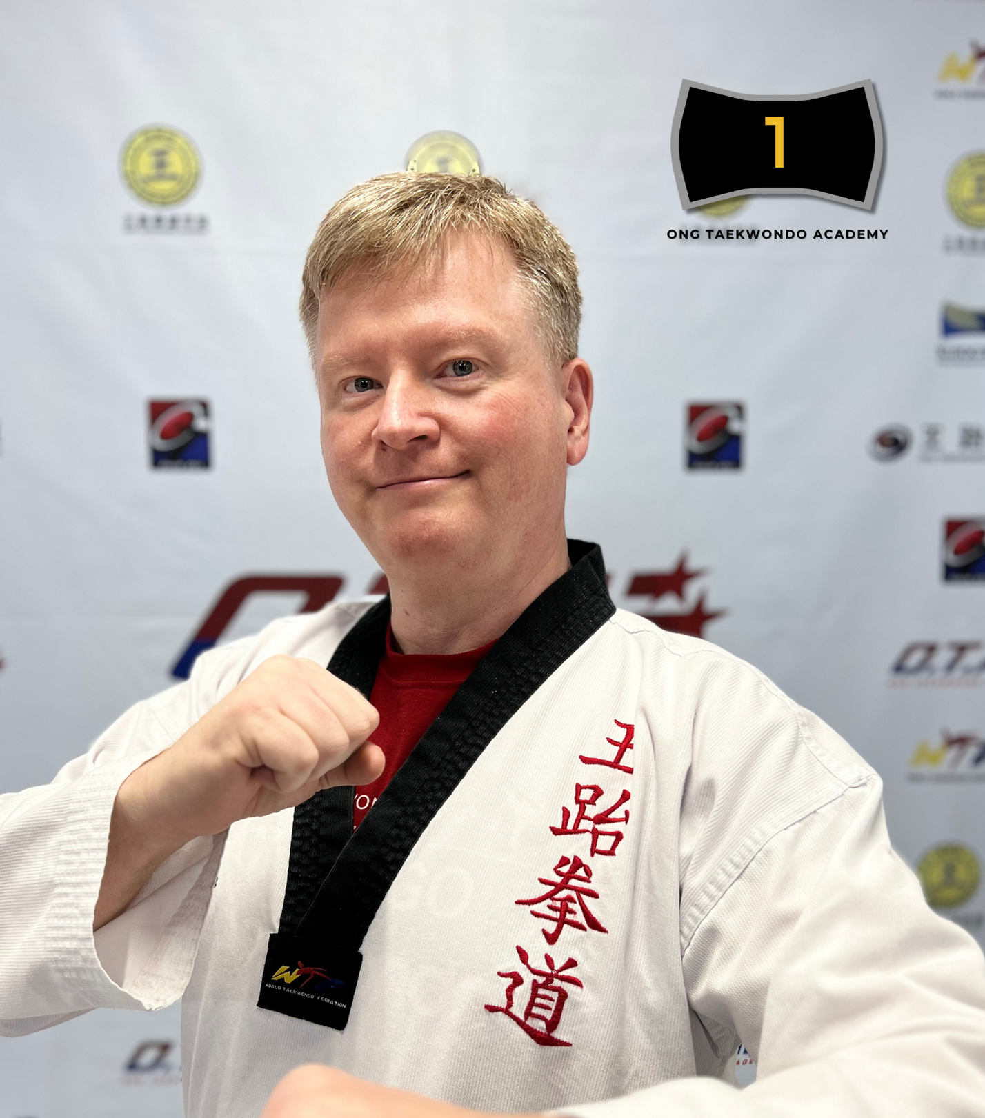 A man in a martial arts uniform standing in front of a banner, showing a fist in a fighting pose. The uniform has Chinese characters embroidered on it. The text overlay at the bottom states: 'Chris VanAllman Black Belt Year 2022'.