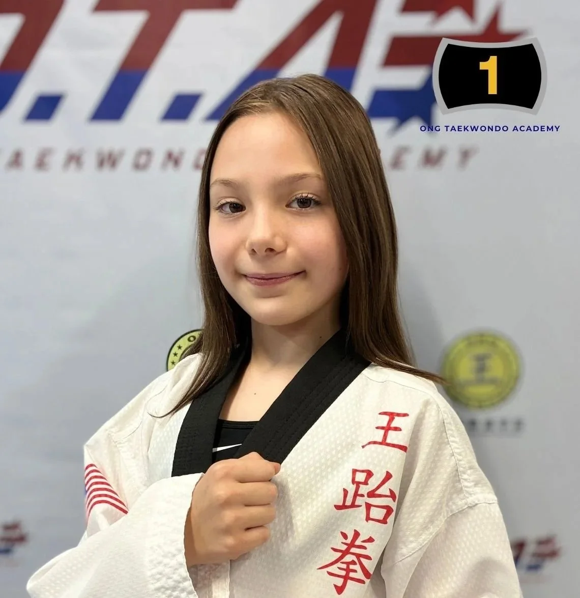 Young girl in a white Taekwondo uniform with red Chinese characters, holding a black belt, standing in front of a martial arts training center background.