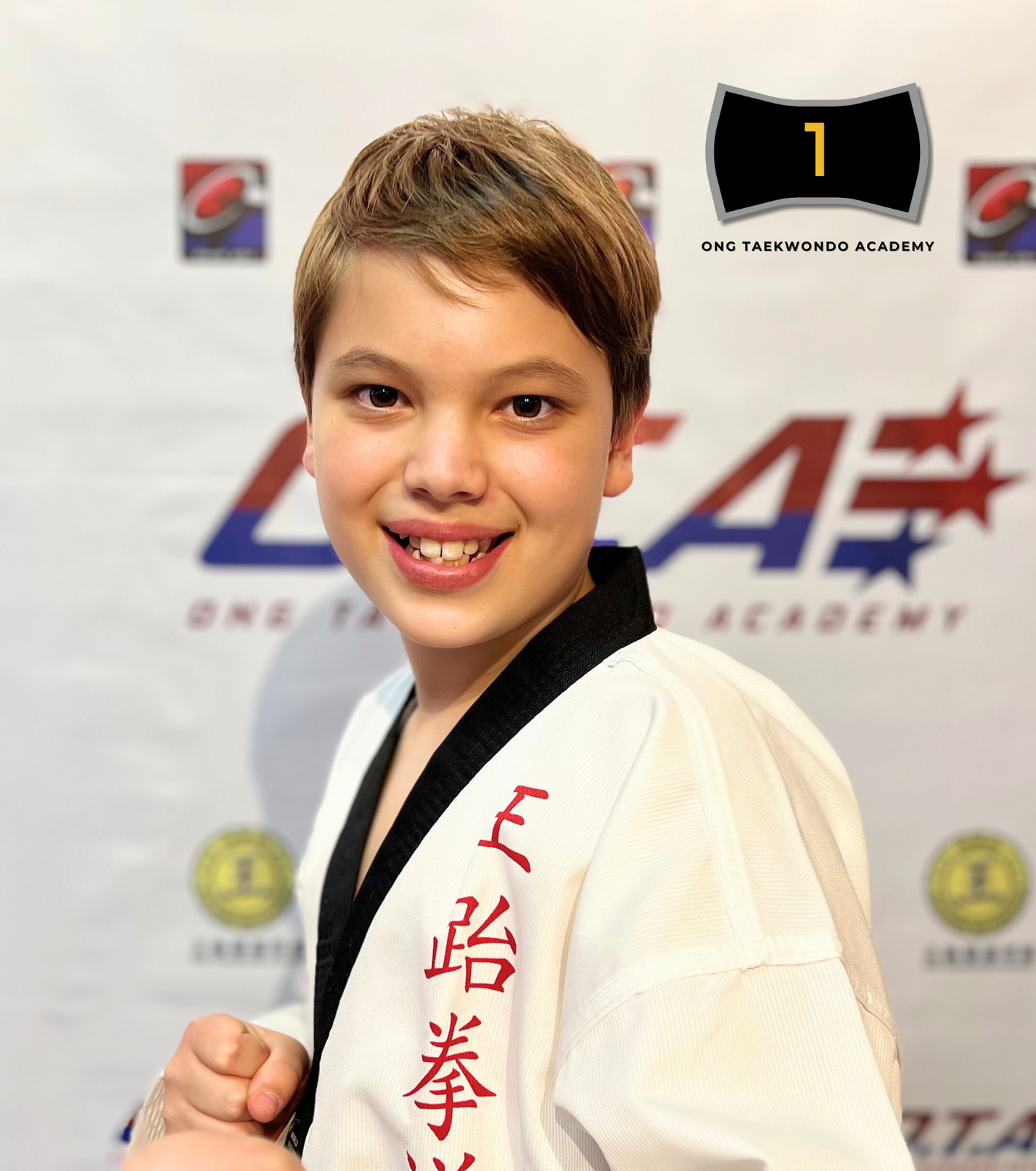 Young boy Kevin McCue in a taekwondo uniform with a black belt, smiling and posing with a fist, in front of a backdrop with logos for a taekwondo academy and other sponsors, celebrating his achievement of a black belt in 2024.