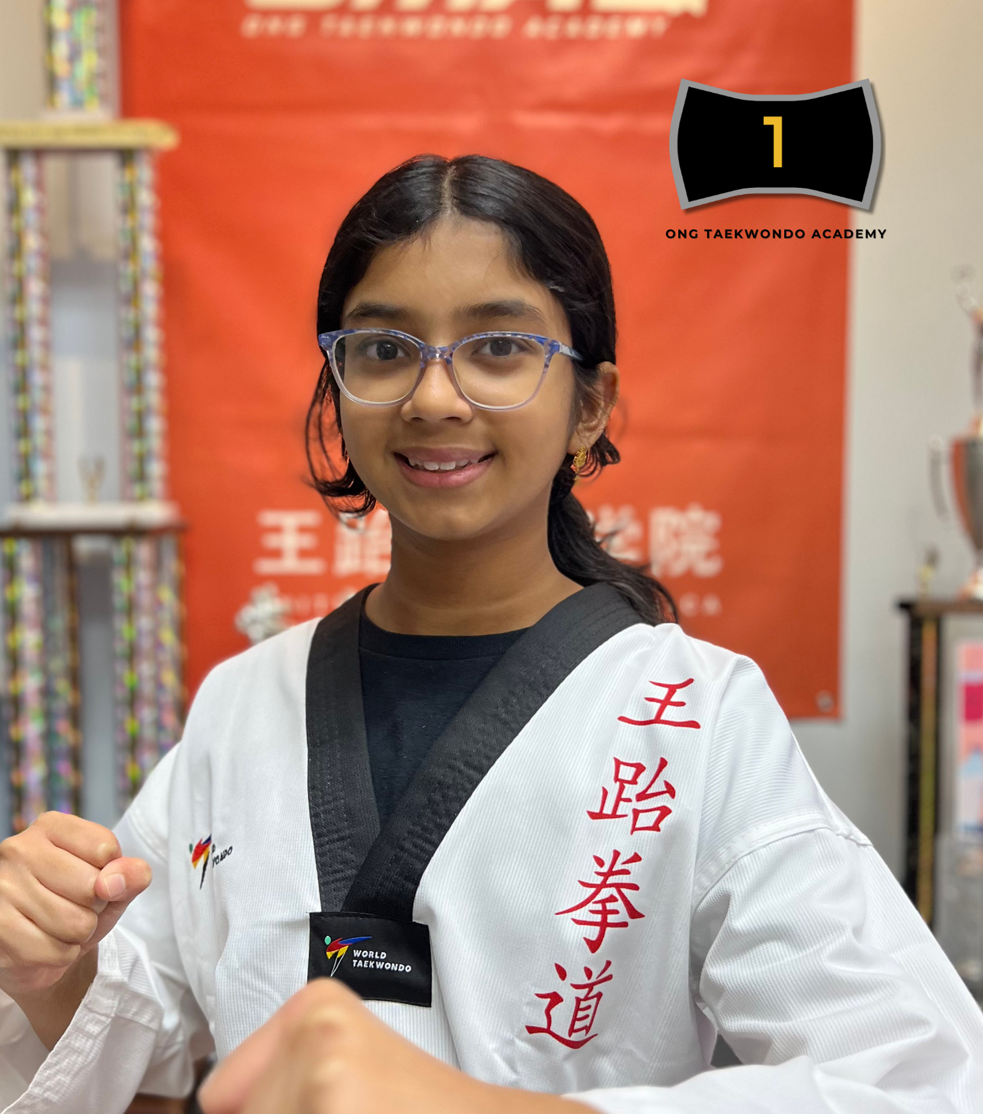 A young girl with glasses smiling and wearing a white taekwondo uniform with red Chinese characters on the side, and a black belt. She is in a martial arts studio with trophies and a red banner in the background, celebrating her ranking in taekwondo for the year 2024.