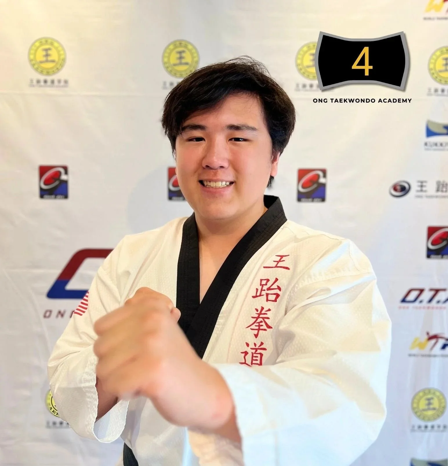 Young man in a taekwondo uniform making a fist with his right hand, standing in front of a backdrop with logos, smiling at the camera.